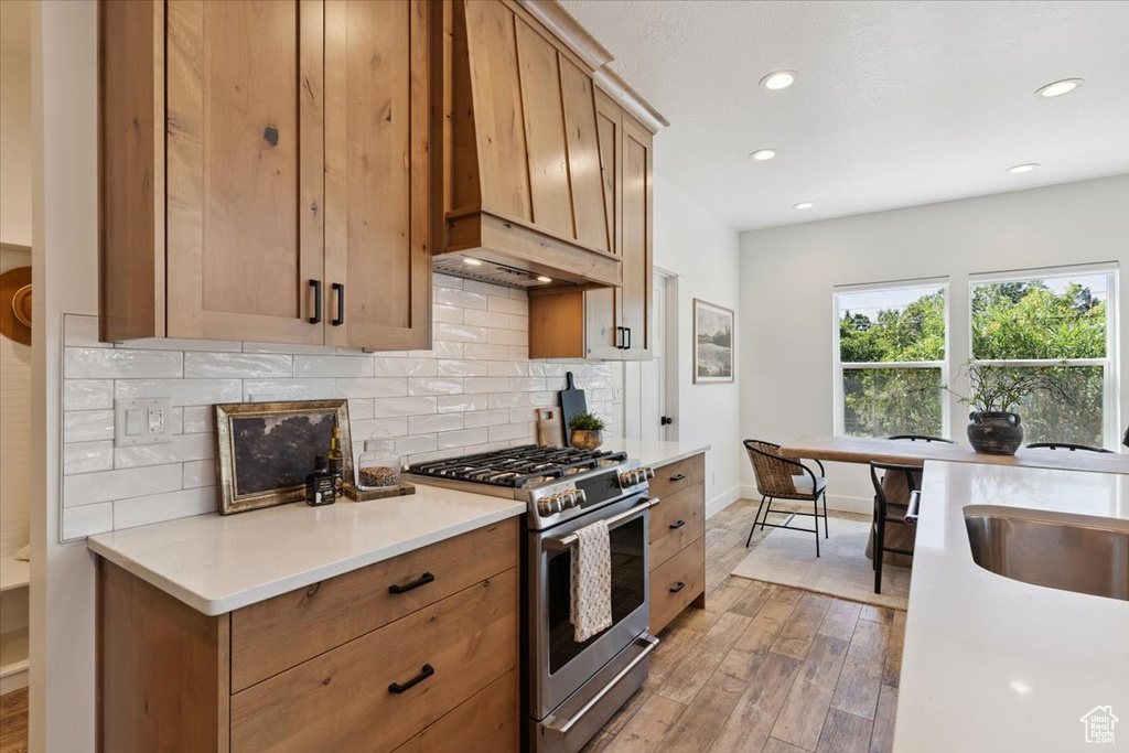 Kitchen featuring stainless steel range with gas cooktop, premium range hood, light wood-style flooring, tasteful backsplash, and light countertops