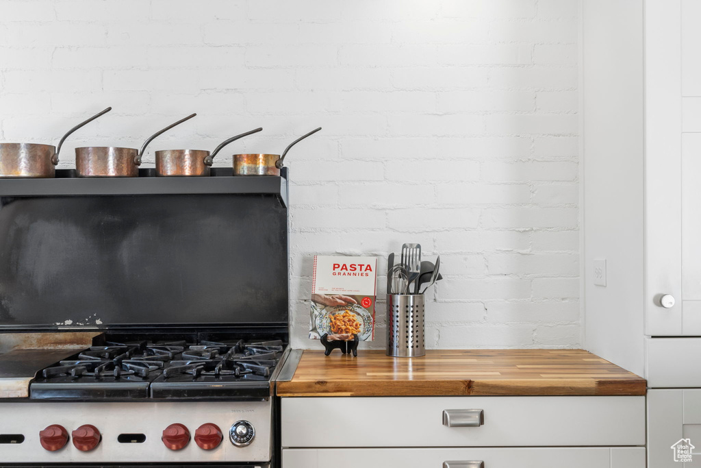 Kitchen with butcher block countertops, stainless steel gas range, white cabinetry, and brick wall
