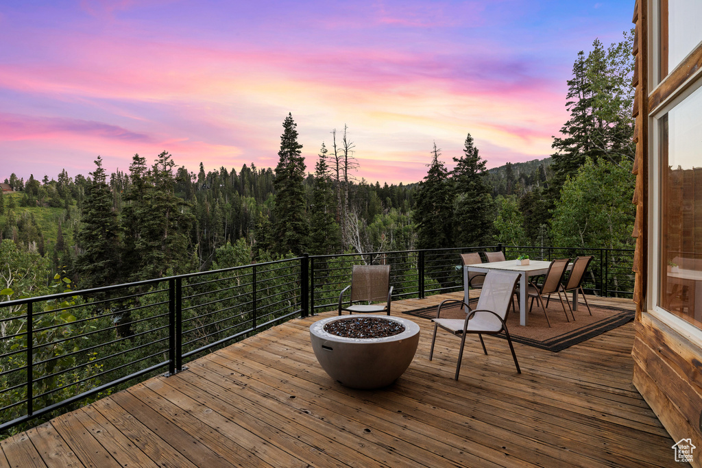 Deck at dusk with a view of trees, outdoor dining space, and a fire pit