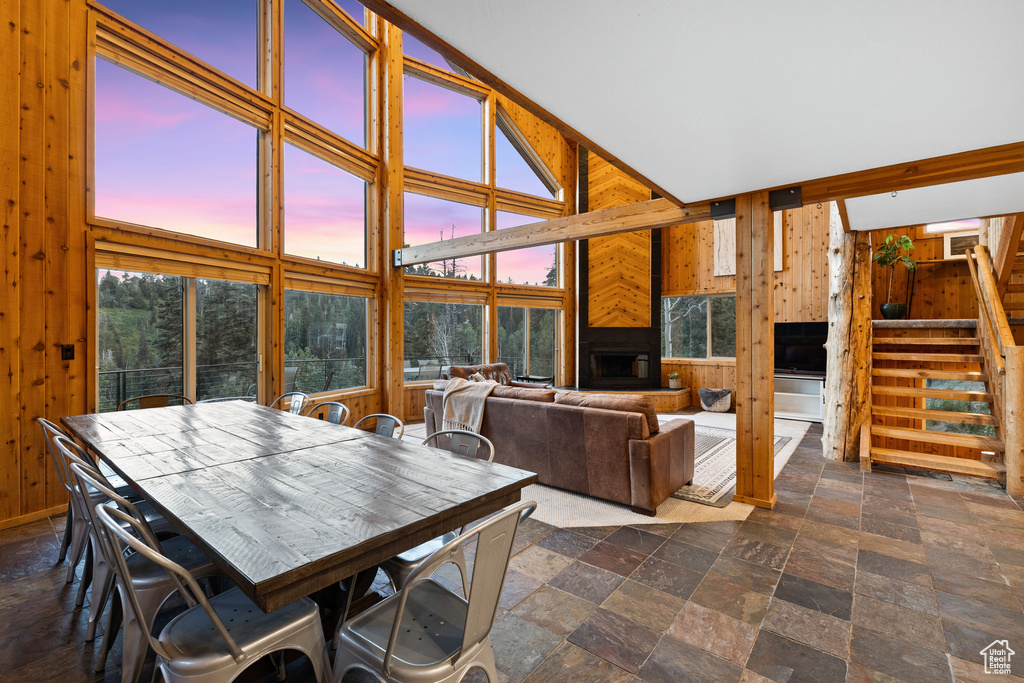 Dining space with wood walls, a fireplace with raised hearth, stairway, dark stone finish flooring, and a towering ceiling