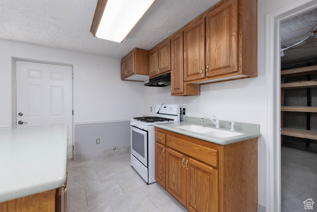 Kitchen featuring white range with gas stovetop, brown cabinetry, light countertops, a textured ceiling, and under cabinet range hood