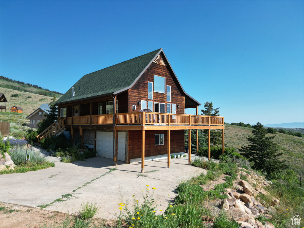 Rear view of house featuring driveway, an attached garage, stairs, a wooden deck, and roof with shingles