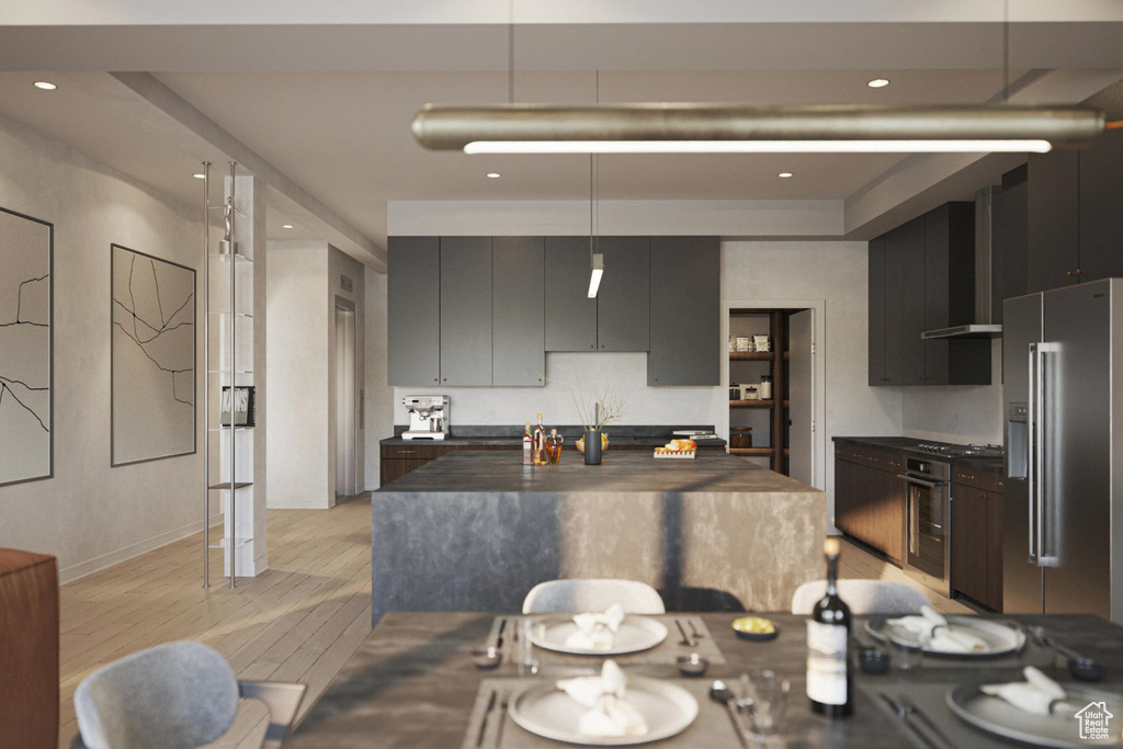 Kitchen featuring dark countertops, stainless steel appliances, light wood-style floors, wall chimney range hood, and hanging light fixtures