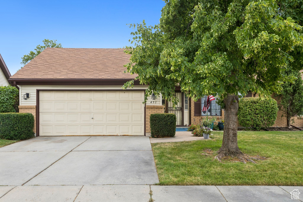 View of property hidden behind natural elements featuring brick siding, driveway, a garage, a front lawn, and roof with shingles