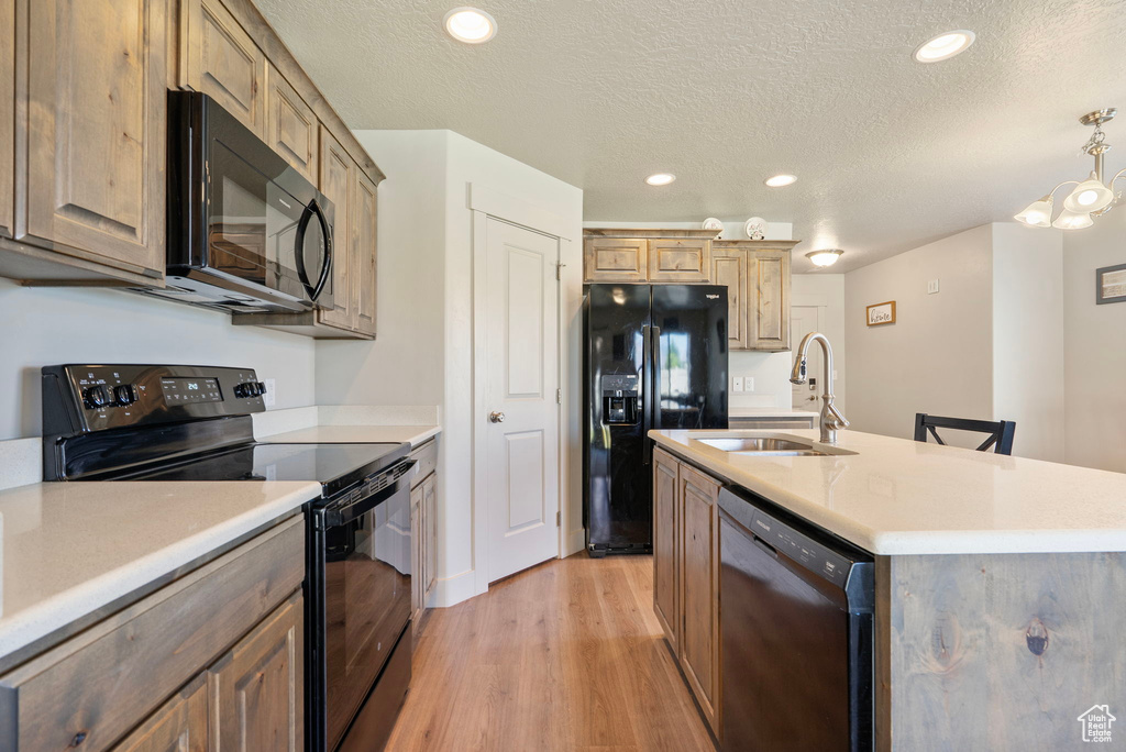 Kitchen with black appliances, light wood-style floors, a textured ceiling, an island with sink, and light countertops