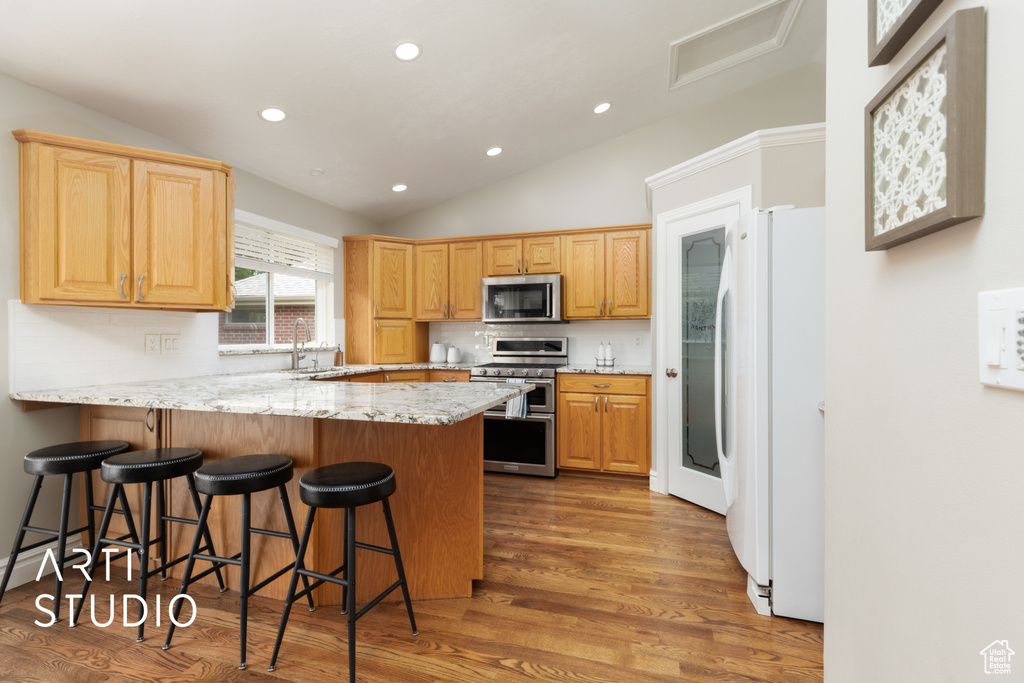 Kitchen with a peninsula, appliances with stainless steel finishes, vaulted ceiling, light wood-style floors, and light stone countertops