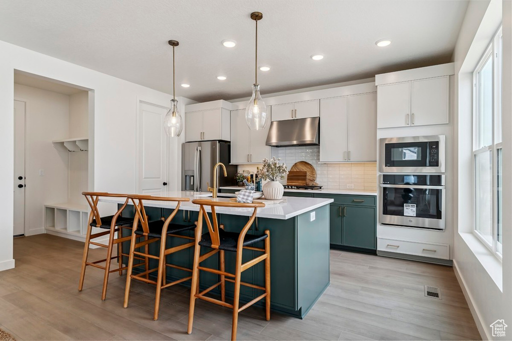 Kitchen with appliances with stainless steel finishes, tasteful backsplash, a center island with sink, light wood-style flooring, and under cabinet range hood