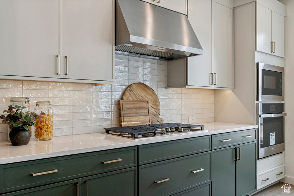 Kitchen with green cabinetry, exhaust hood, stainless steel appliances, and backsplash