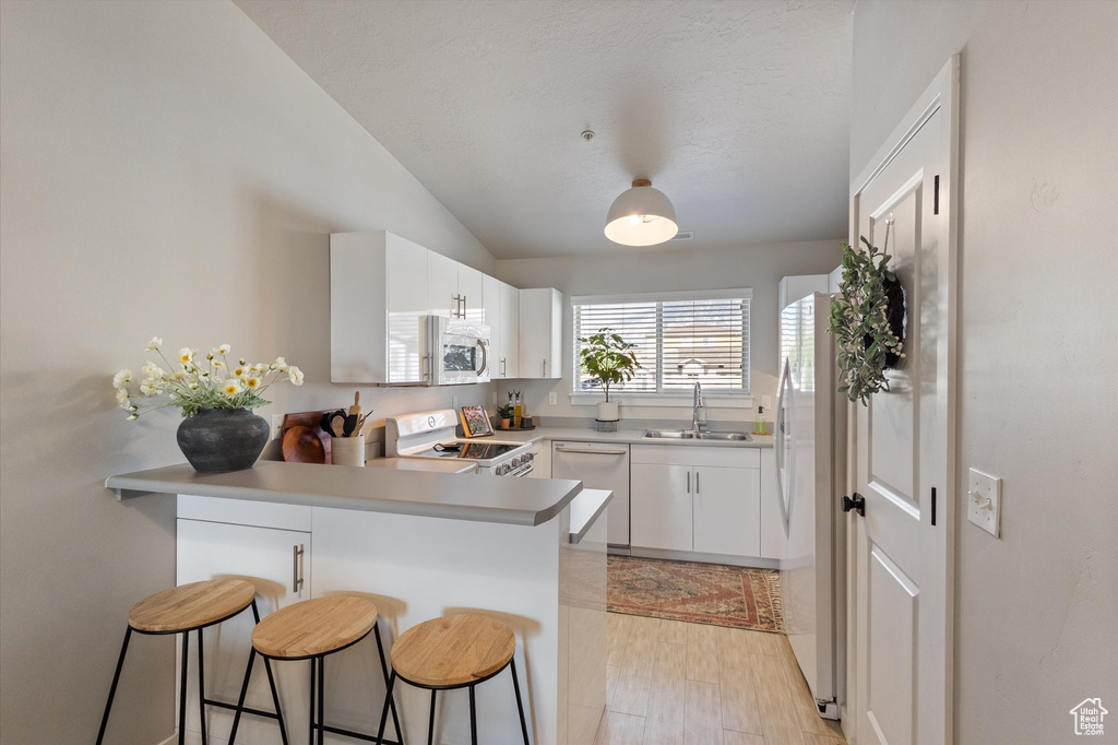 Kitchen featuring a peninsula, white appliances, a kitchen bar, lofted ceiling, and white cabinetry