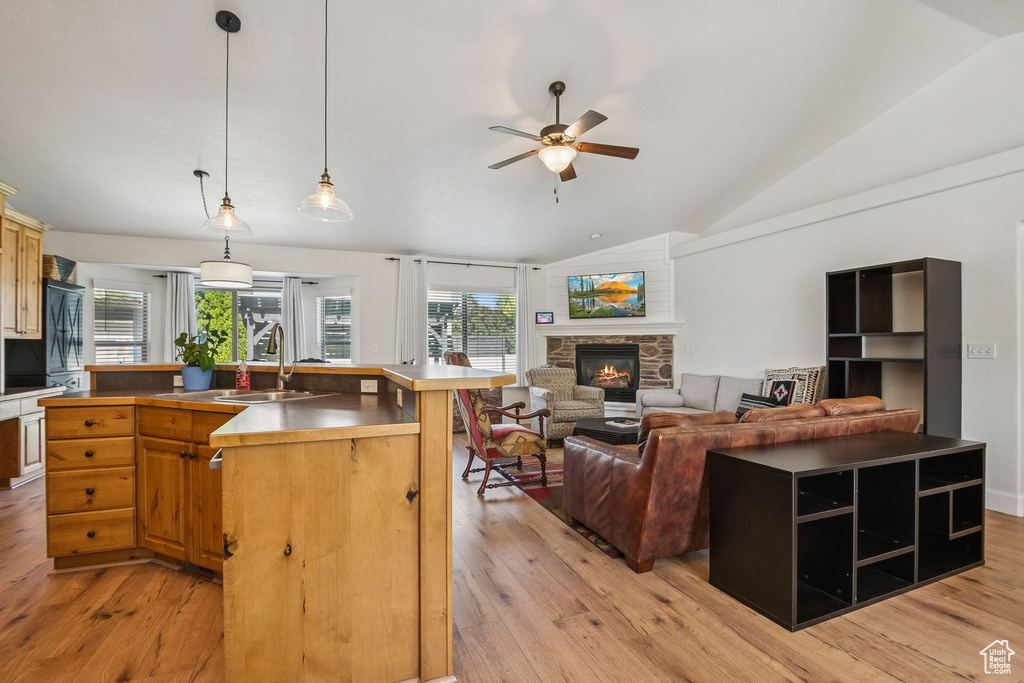 Kitchen with vaulted ceiling, a stone fireplace, a center island with sink, ceiling fan, and light wood-style floors