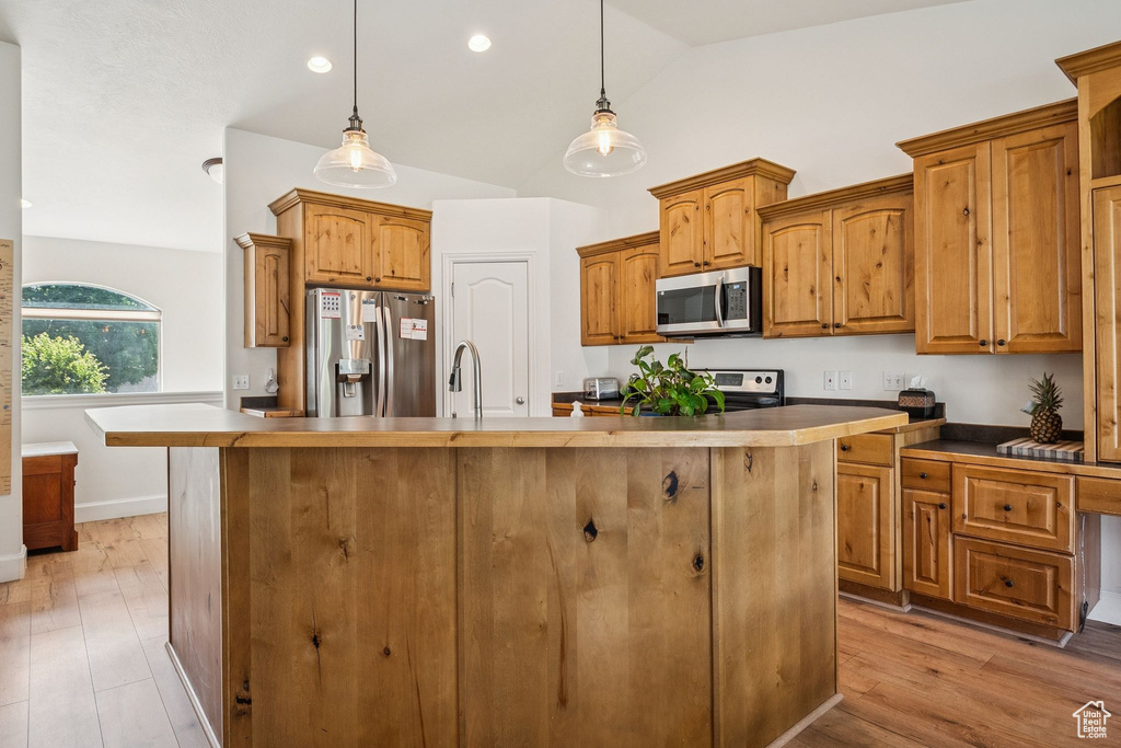 Kitchen with appliances with stainless steel finishes, lofted ceiling, an island with sink, brown cabinetry, and decorative light fixtures