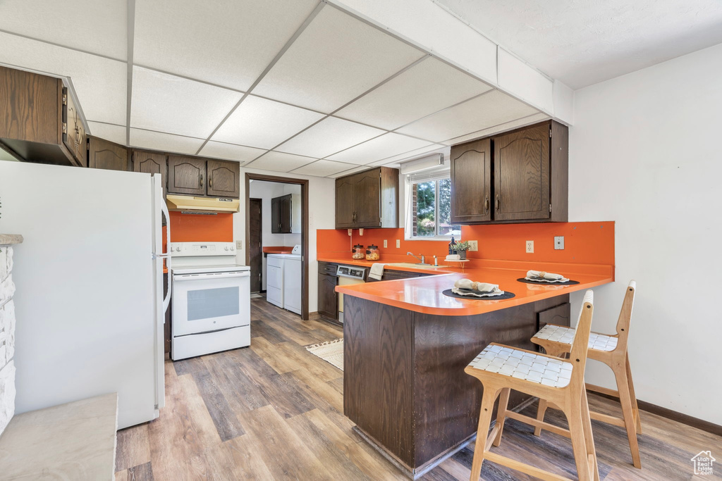 Kitchen featuring white appliances, light wood-style flooring, dark brown cabinets, a peninsula, and a breakfast bar