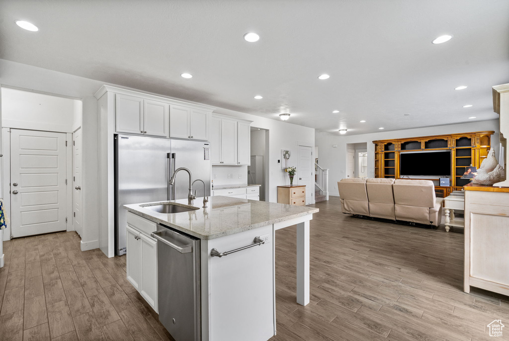 Kitchen featuring a kitchen island with sink, stainless steel appliances, wood finished floors, white cabinetry, and light stone counters
