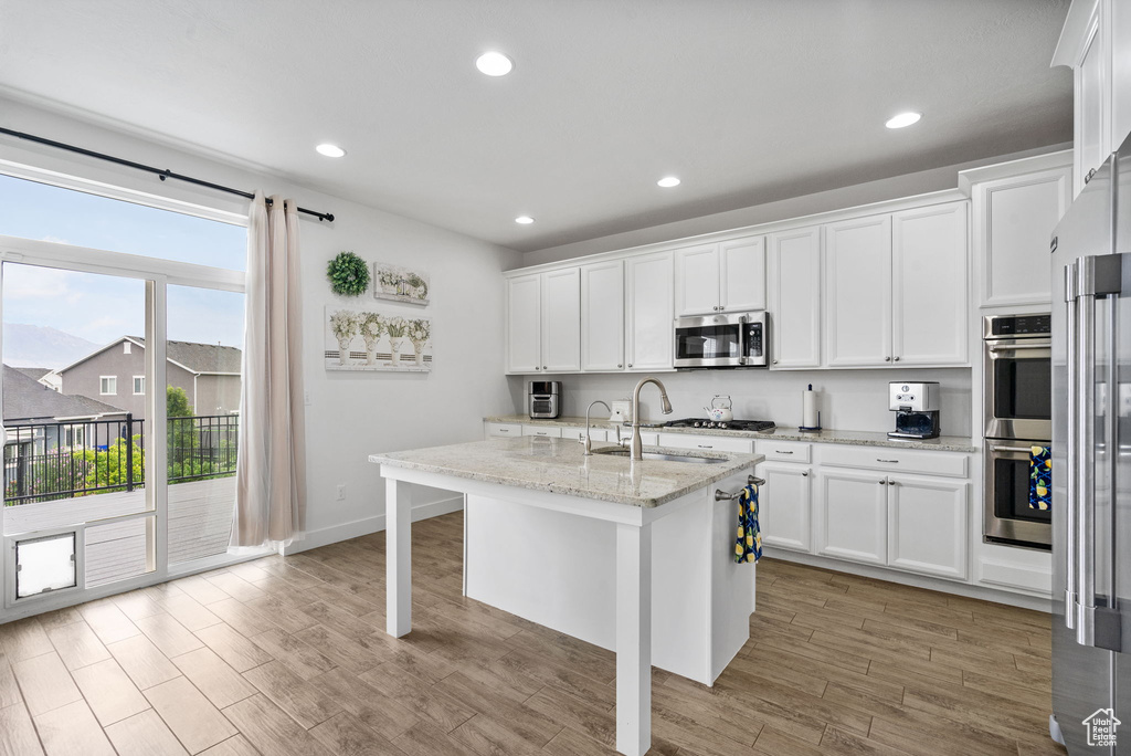 Kitchen featuring white cabinets, appliances with stainless steel finishes, light stone countertops, light wood-style floors, and a kitchen island with sink