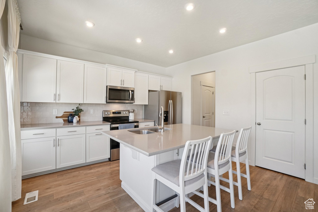 Kitchen with appliances with stainless steel finishes, a kitchen island with sink, decorative backsplash, light wood-style floors, and a kitchen breakfast bar