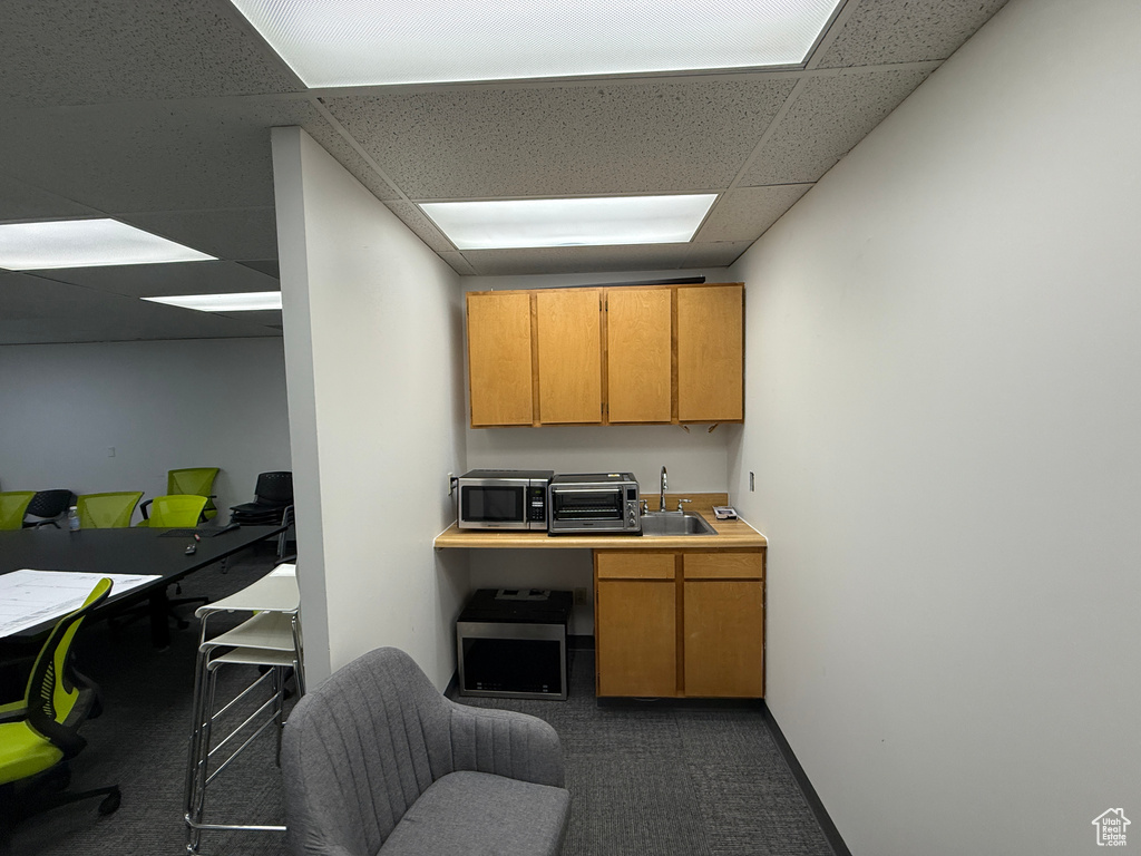 Kitchen featuring dark colored carpet, a drop ceiling, stainless steel microwave, a desk, and brown cabinets