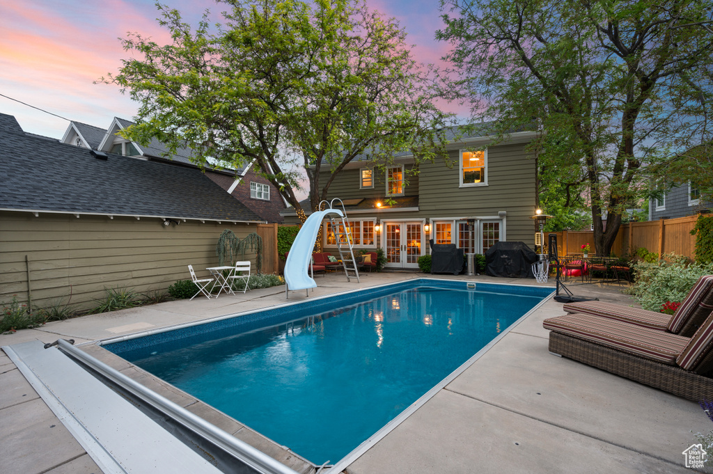 Pool at dusk with french doors, a water slide, a fenced backyard, a patio, and area for grilling