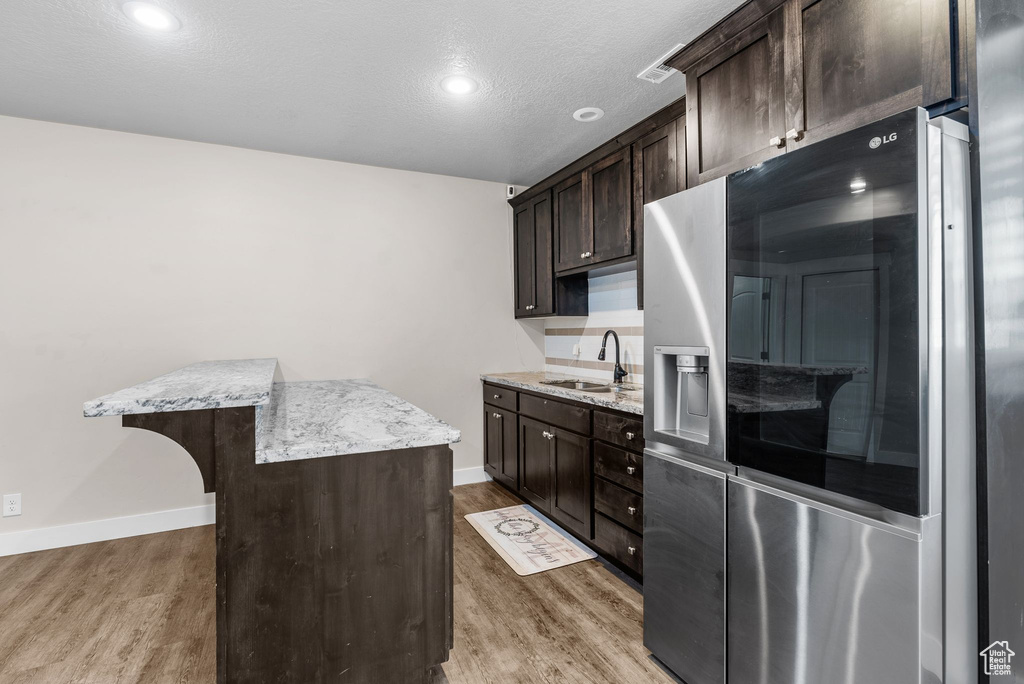 Kitchen with stainless steel fridge with ice dispenser, dark brown cabinetry, wood finished floors, light stone countertops, and a textured ceiling