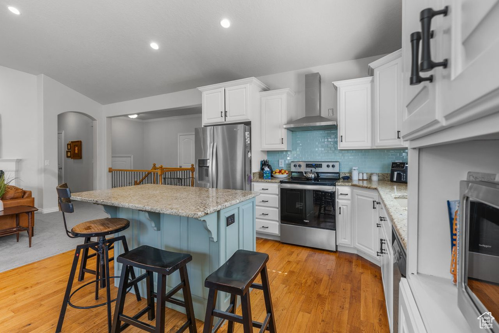 Kitchen featuring appliances with stainless steel finishes, wall chimney exhaust hood, white cabinets, a breakfast bar, and a center island
