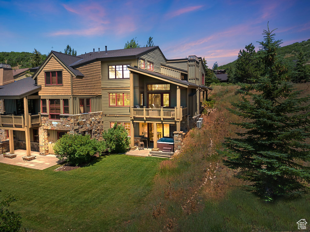 Back of house at dusk with a patio area, stone siding, a lawn, and a balcony
