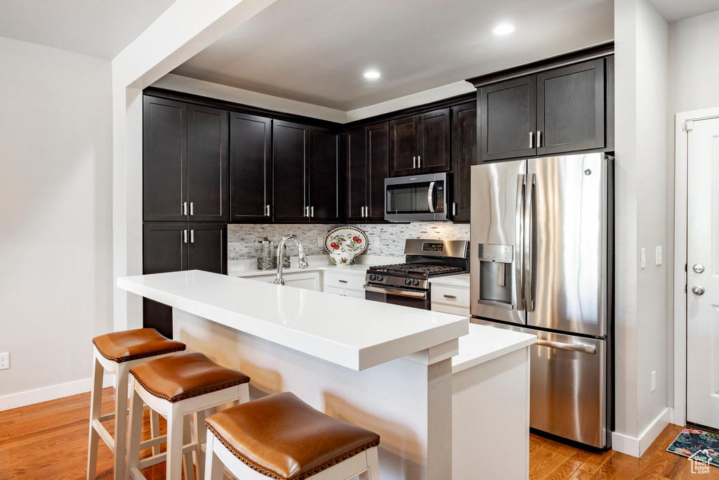 Kitchen with appliances with stainless steel finishes, a breakfast bar, backsplash, light wood finished floors, and light countertops