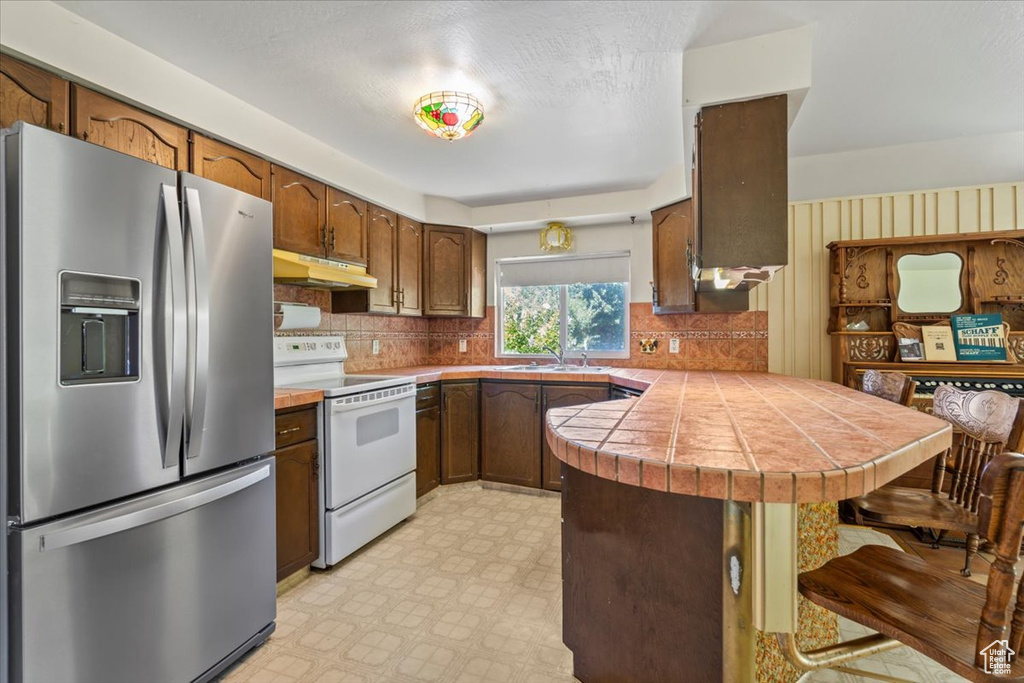 Kitchen featuring stainless steel fridge, a peninsula, white range with electric stovetop, a breakfast bar, and light flooring
