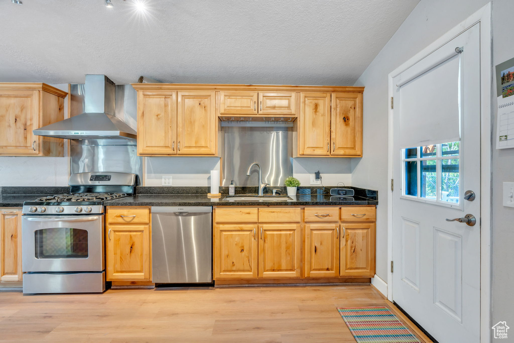 Kitchen with appliances with stainless steel finishes, wall chimney exhaust hood, light wood finished floors, a textured ceiling, and dark stone countertops