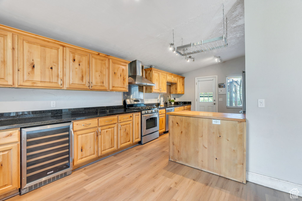Kitchen with light wood finished floors, beverage cooler, appliances with stainless steel finishes, vaulted ceiling, and wall chimney exhaust hood