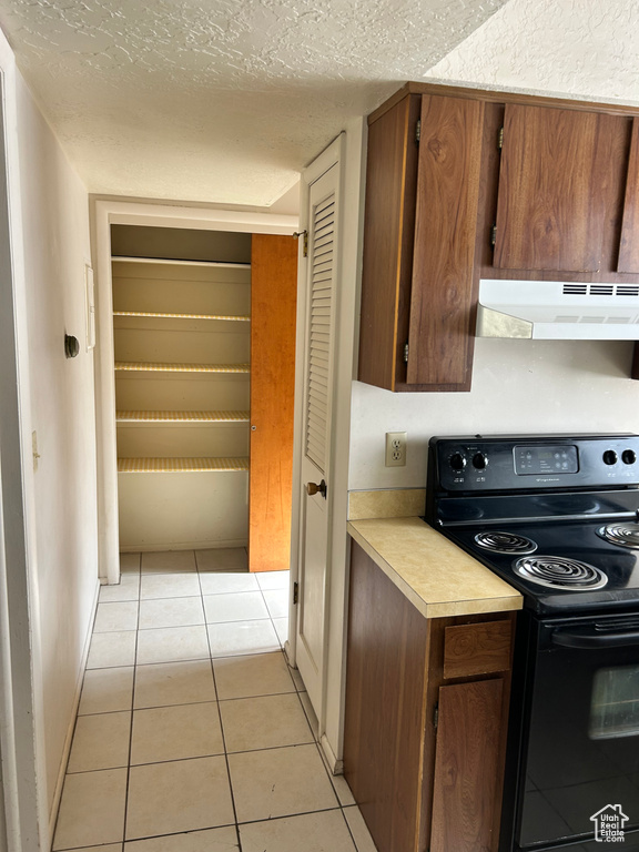 Kitchen featuring black electric range, light countertops, under cabinet range hood, light tile patterned floors, and a textured ceiling