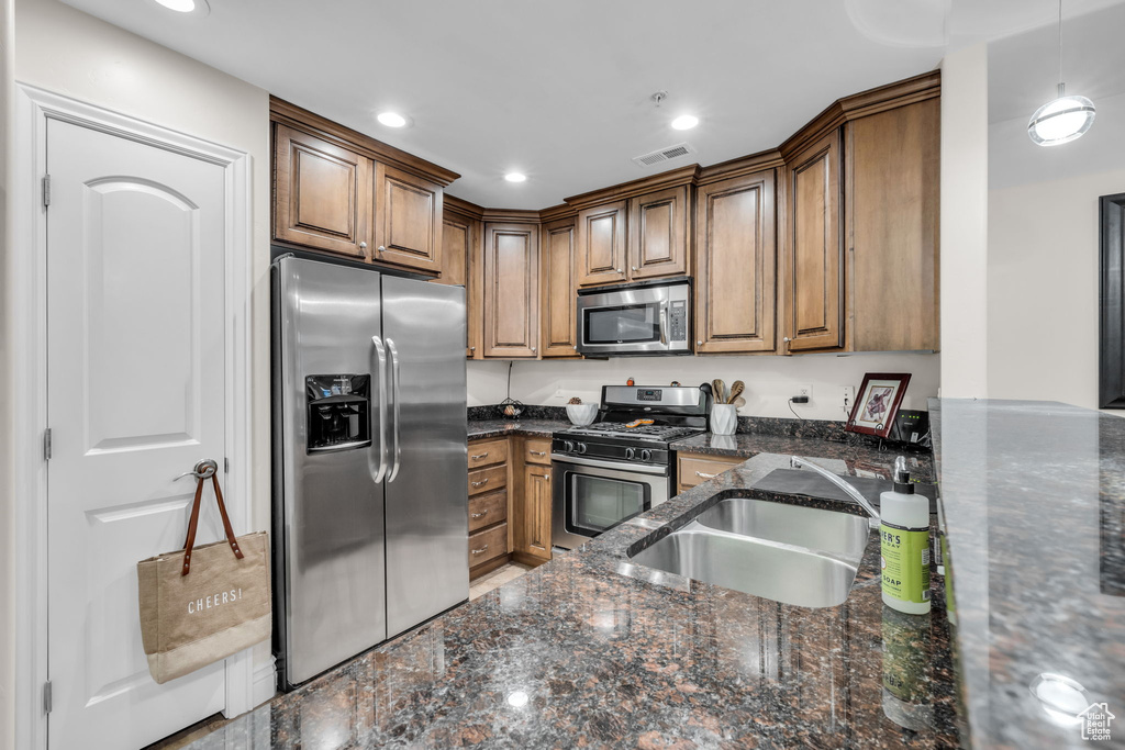 Kitchen with appliances with stainless steel finishes, dark stone countertops, brown cabinetry, hanging light fixtures, and recessed lighting