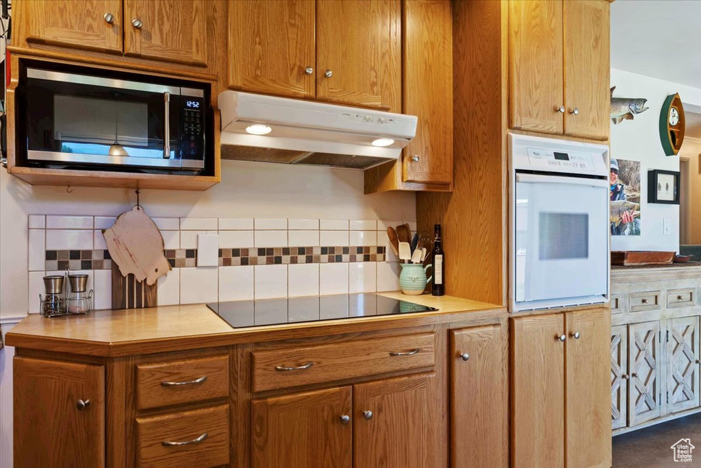 Kitchen featuring white oven, tasteful backsplash, under cabinet range hood, stainless steel microwave, and brown cabinetry
