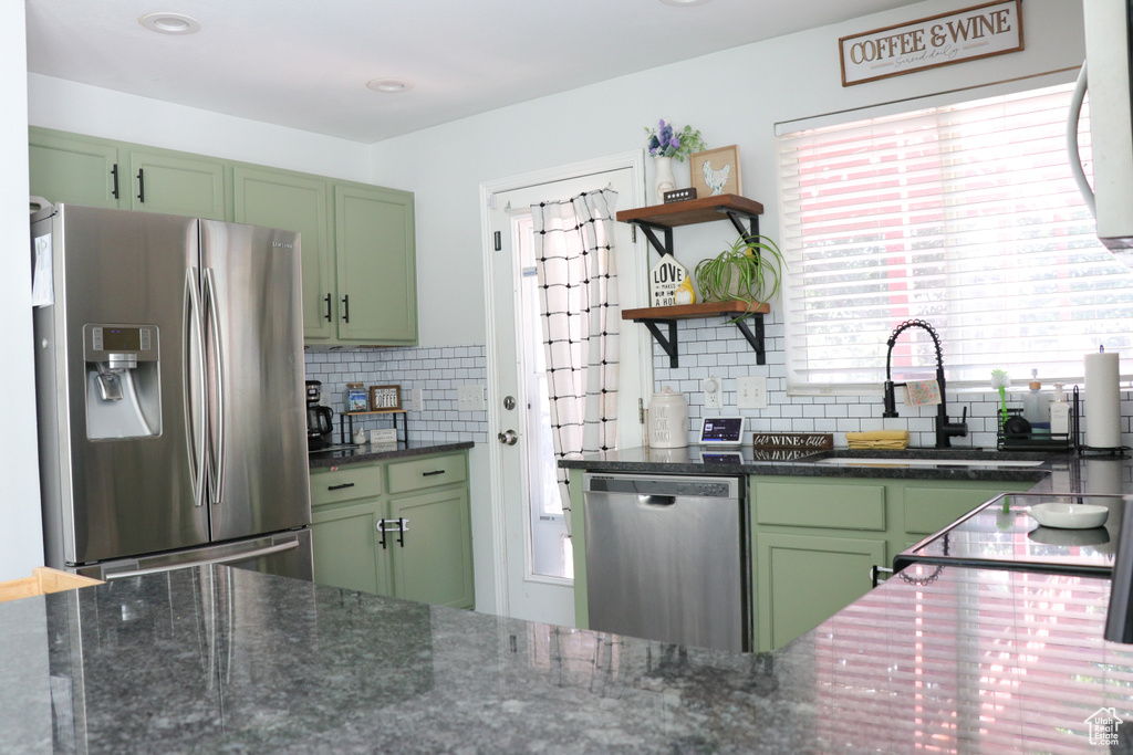 Kitchen with green cabinets, appliances with stainless steel finishes, decorative backsplash, open shelves, and dark stone countertops