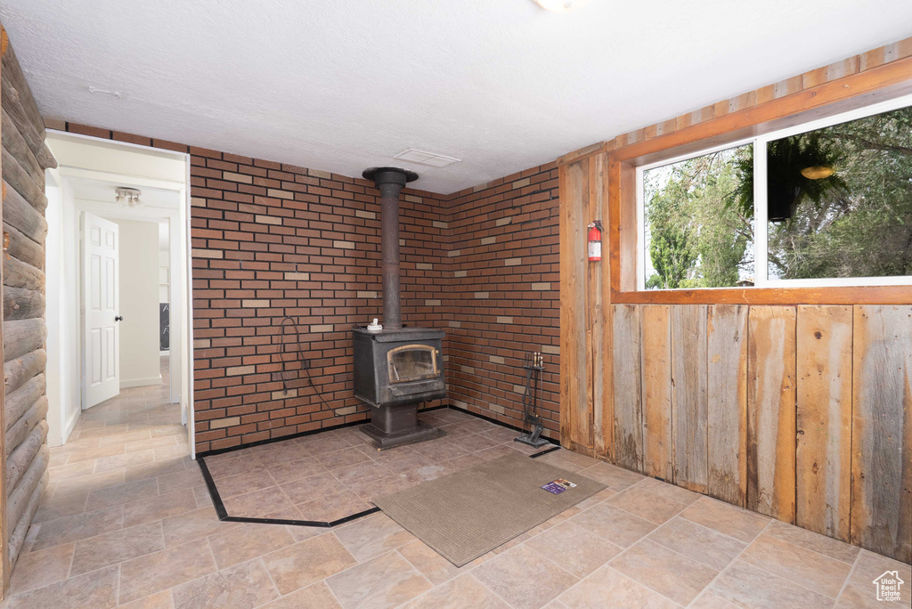 Unfurnished living room with a wood stove, light stone finish floors, wood walls, and brick wall