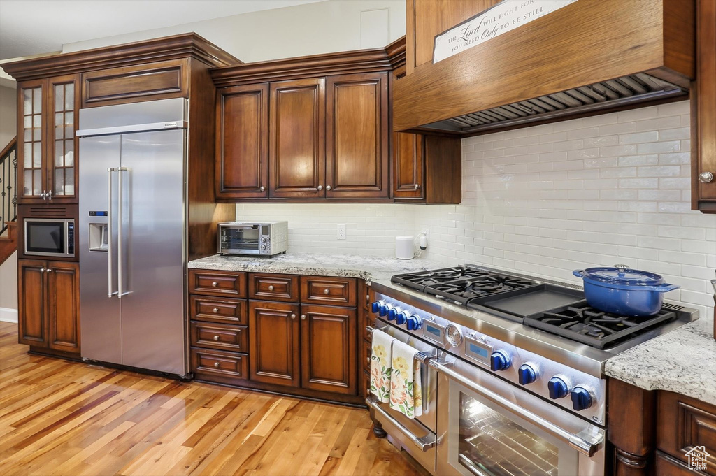 Kitchen with built in appliances, backsplash, custom exhaust hood, light wood-type flooring, and light stone countertops