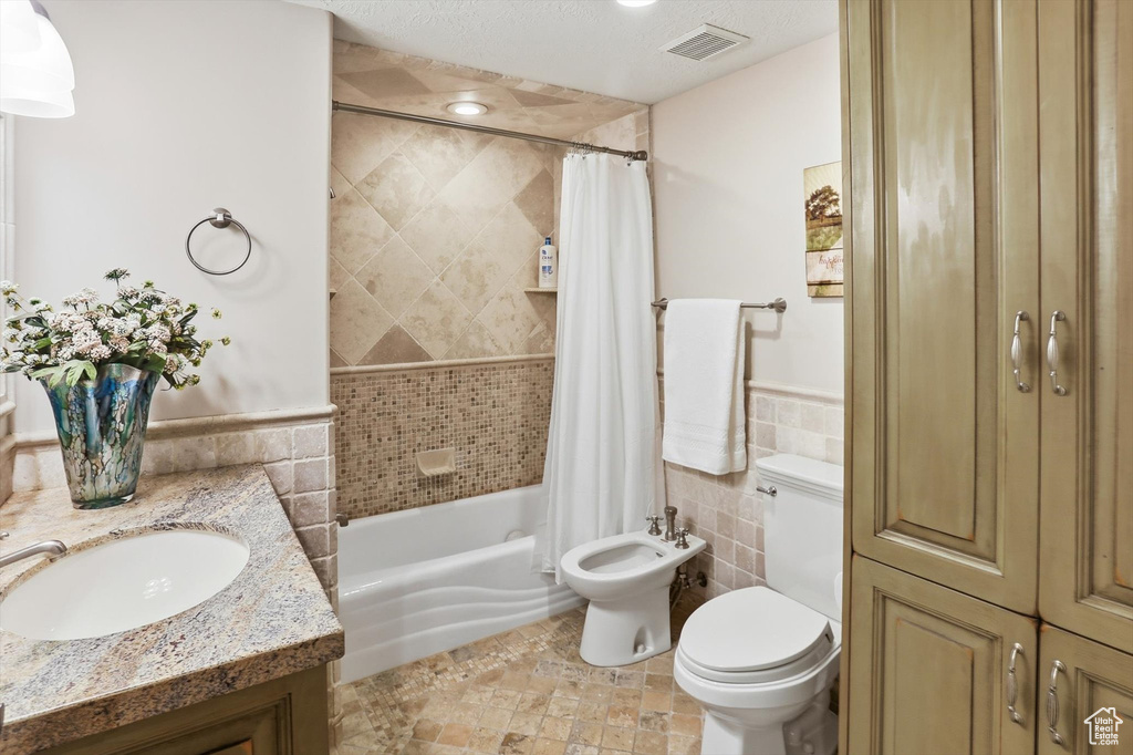 Bathroom featuring tile walls, shower / bath combination with curtain, wainscoting, a bidet, and a textured ceiling