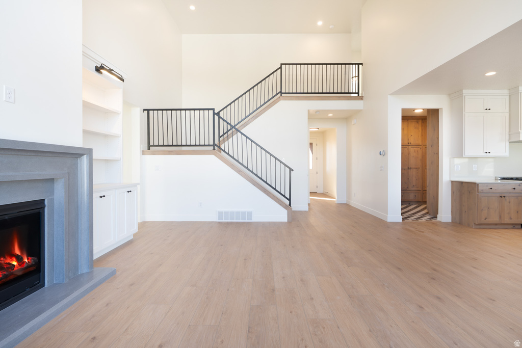 Unfurnished living room featuring a towering ceiling, recessed lighting, light wood finished floors, a warm lit fireplace, and stairway