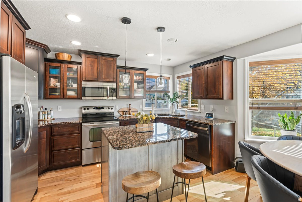 Kitchen featuring appliances with stainless steel finishes, dark stone countertops, light wood finished floors, glass insert cabinets, and hanging light fixtures