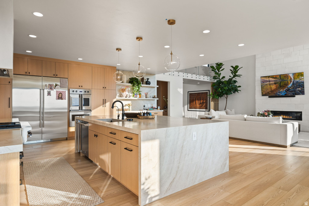Kitchen with a large fireplace, light brown cabinetry, appliances with stainless steel finishes, hanging light fixtures, and an island with sink