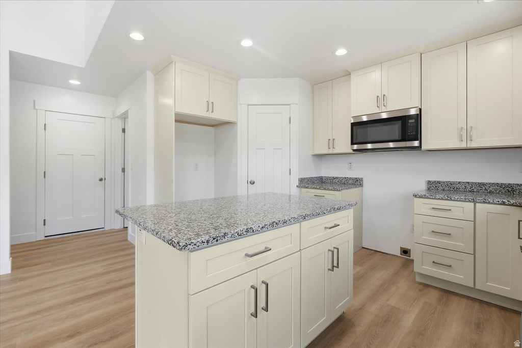 Modern kitchen featuring an island with a speckled countertop, light wood-finish flooring, recessed lighting, white shaker-style cabinetry, and a stainless steel microwave