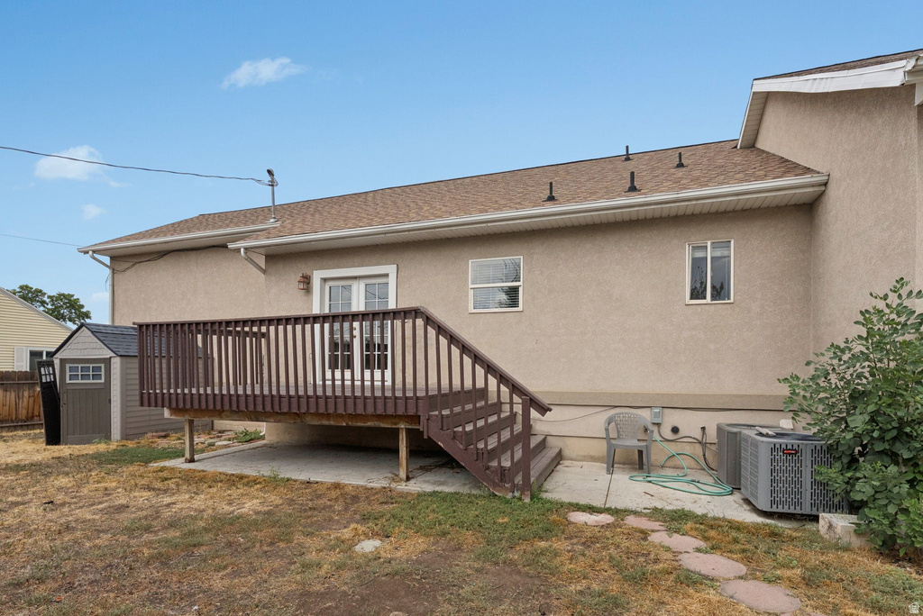 Back of house featuring a deck, stucco siding, a storage shed, a patio area, and roof with shingles