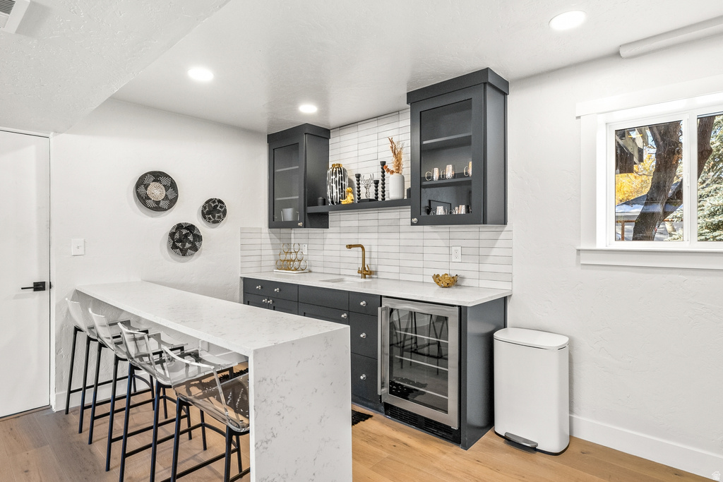 Indoor wet bar featuring wine cooler, light stone countertops, glass fronted cabinets, decorative backsplash, and light wood finished floors