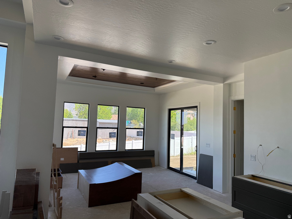 Recessed lighting illuminates the open living space featuring a tray ceiling with a wood-finish accent panel, multiple large windows with black frames, and a glass sliding door providing access to the exterior