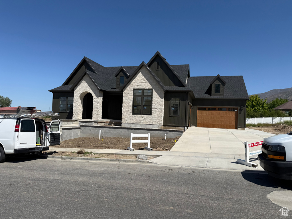 Contemporary residence featuring a dark shingle roof, light-colored stone veneer, dark vertical siding, an arched entryway, and an attached garage with a wood-finish door
