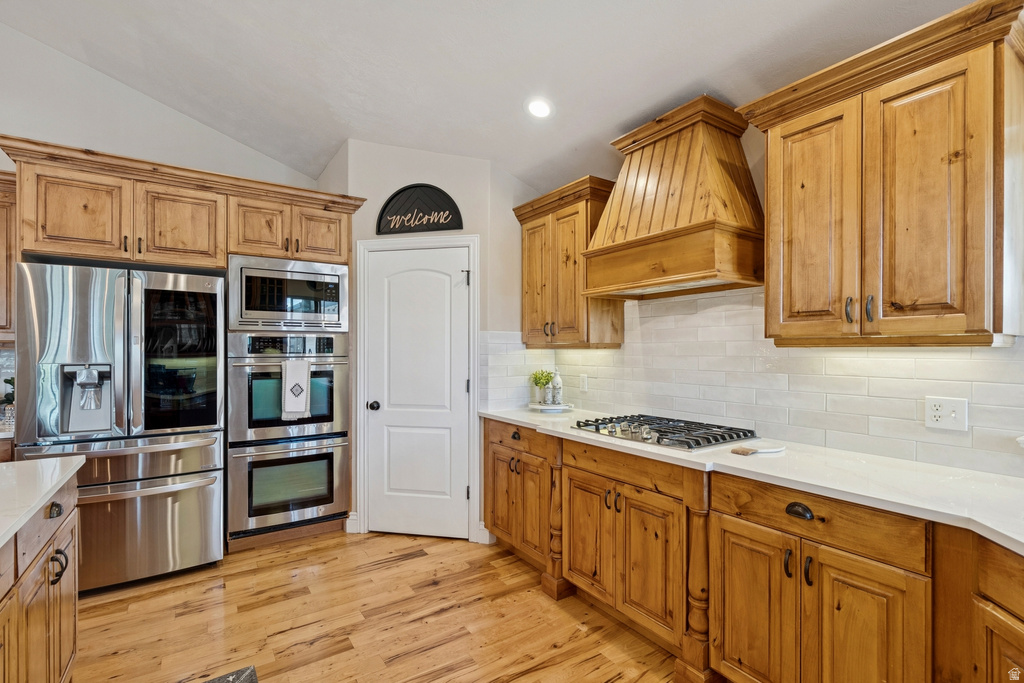 Kitchen featuring stainless steel appliances, wood finish cabinetry, light wood finished floors, light stone countertops, and vaulted ceiling