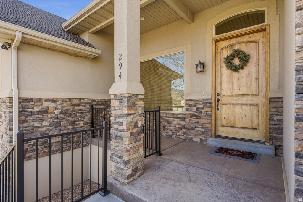 Doorway to property with stone siding, covered porch, stucco siding, and roof with shingles