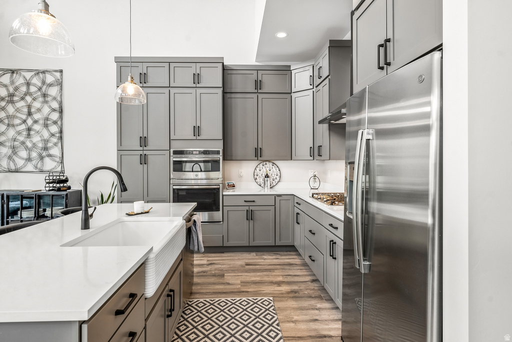 Kitchen with stainless steel appliances, light stone counters, light wood-style floors, pendant lighting, and an island with sink