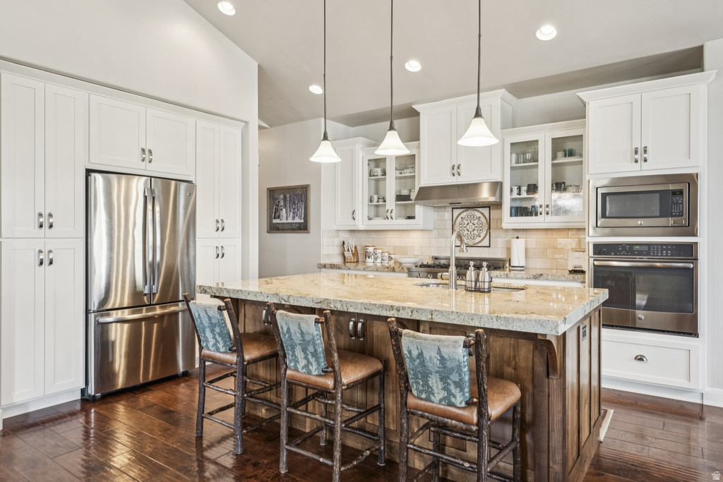 Kitchen featuring stainless steel appliances, light stone countertops, a kitchen island with sink, a breakfast bar area, and glass insert cabinets