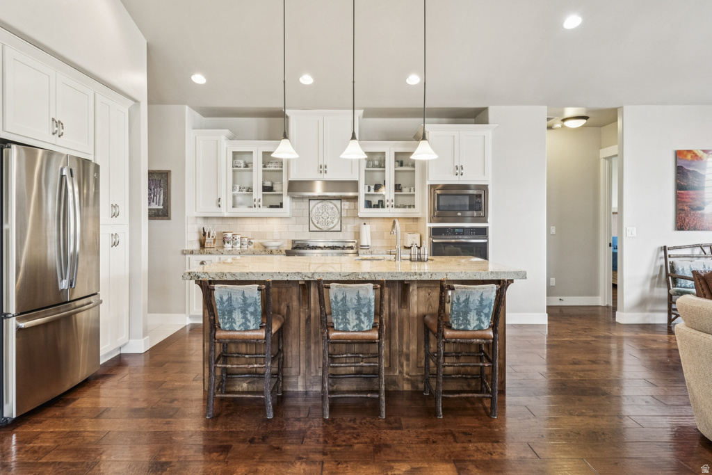 Kitchen featuring glass fronted cabinets, stainless steel appliances, a kitchen island with sink, dark wood finished floors, and a breakfast bar