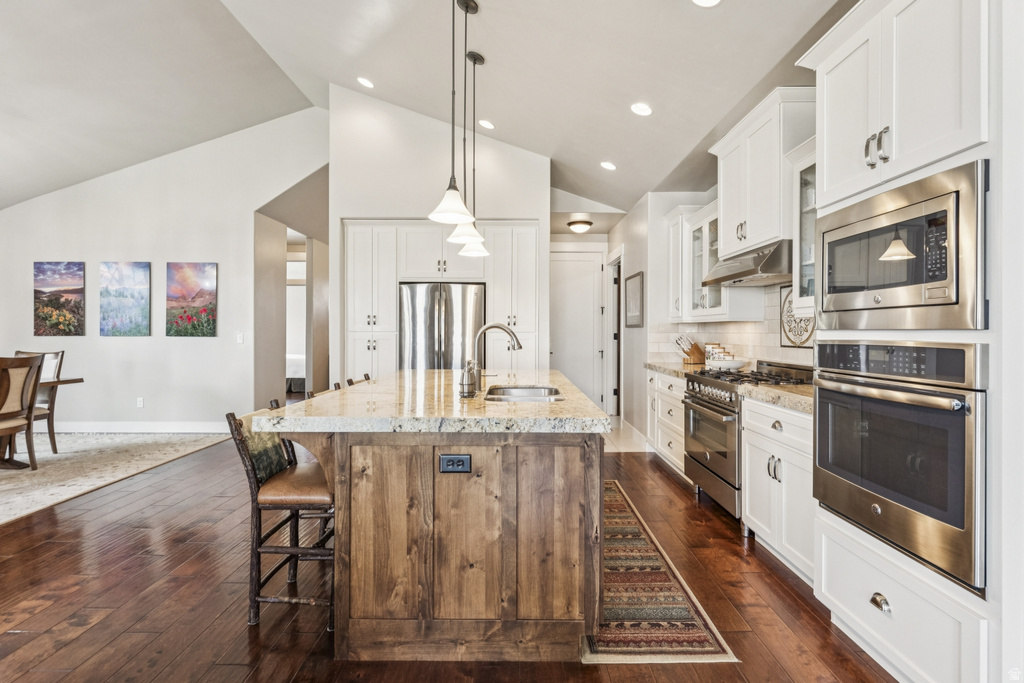 Kitchen with stainless steel appliances, decorative light fixtures, an island with sink, dark wood-style flooring, and light stone counters