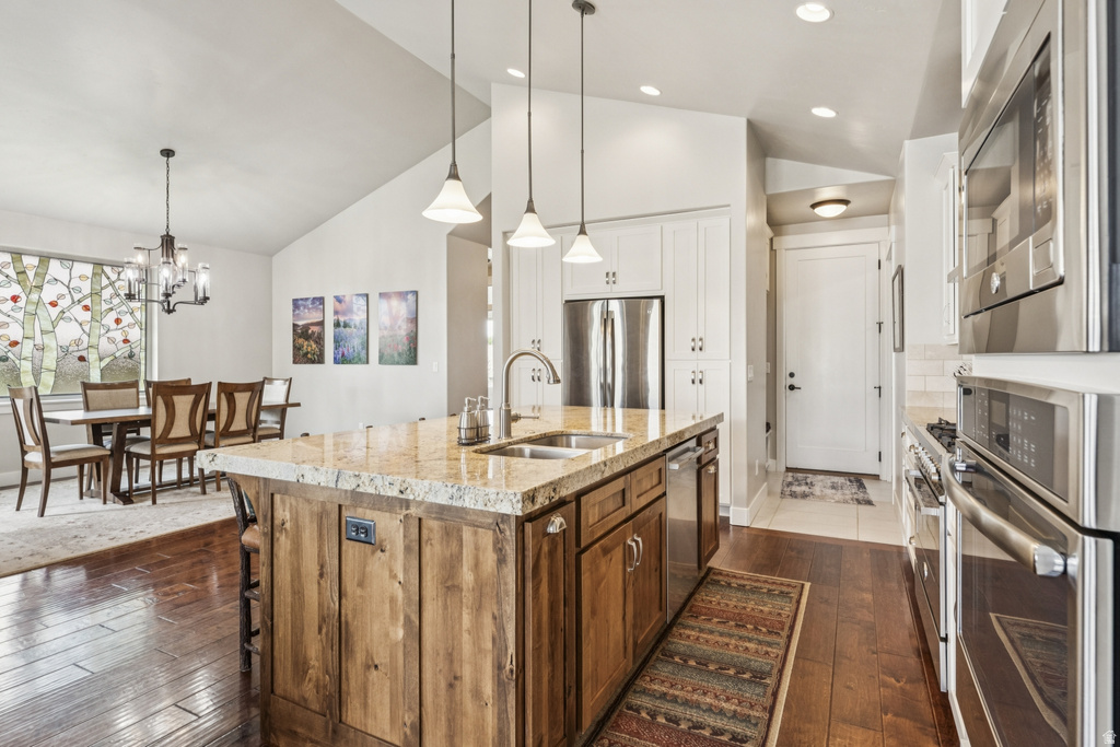 Two tone kitchen with stainless steel appliances, vaulted ceiling, light stone countertops, an island with sink, and two tone cabinetry